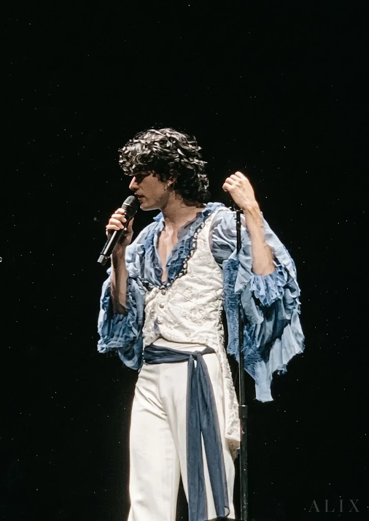 Conan Gray singing into a microphone against a black stage backdrop.