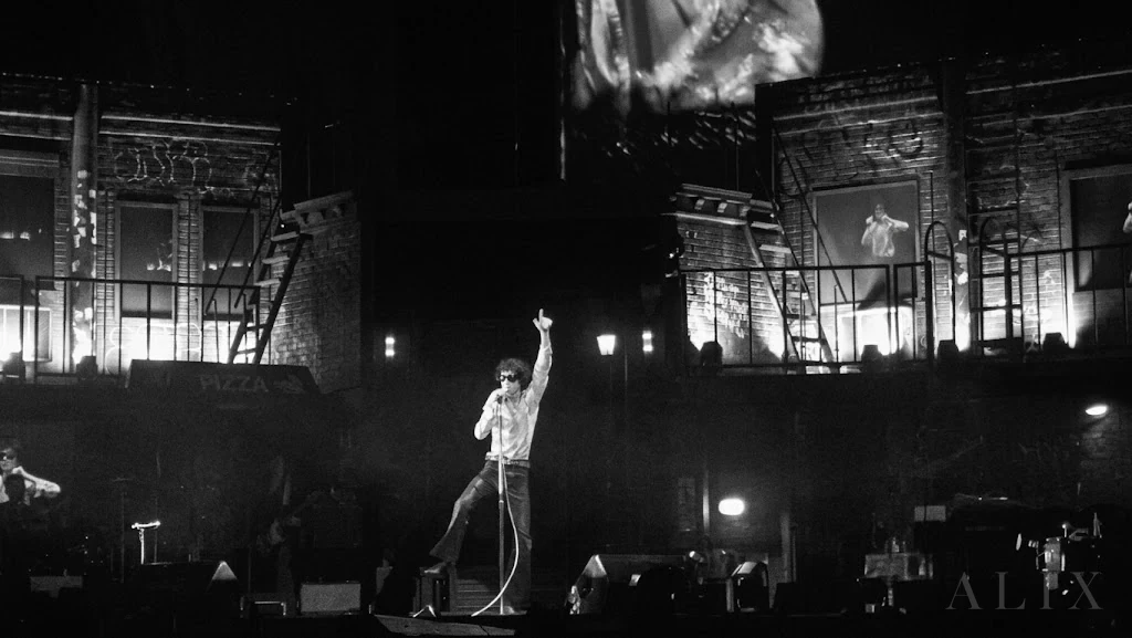 Wide black-and-white photograph of sombr crossing a concert stage above the crowd.