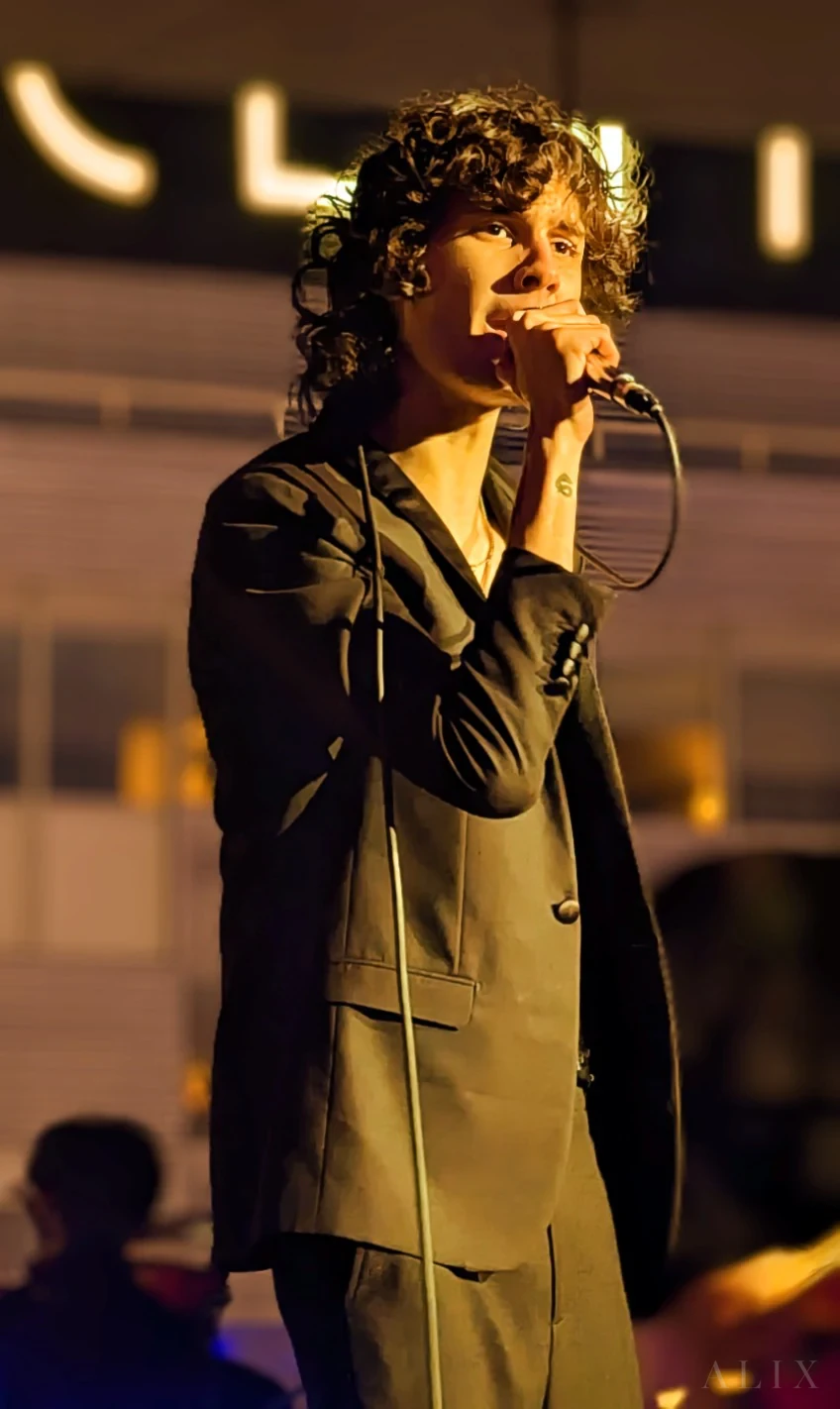 sombr singing into a handheld microphone in warm indoor lighting.