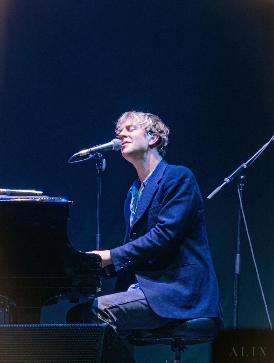Tom Odell sings at a grand piano under blue stage lighting.