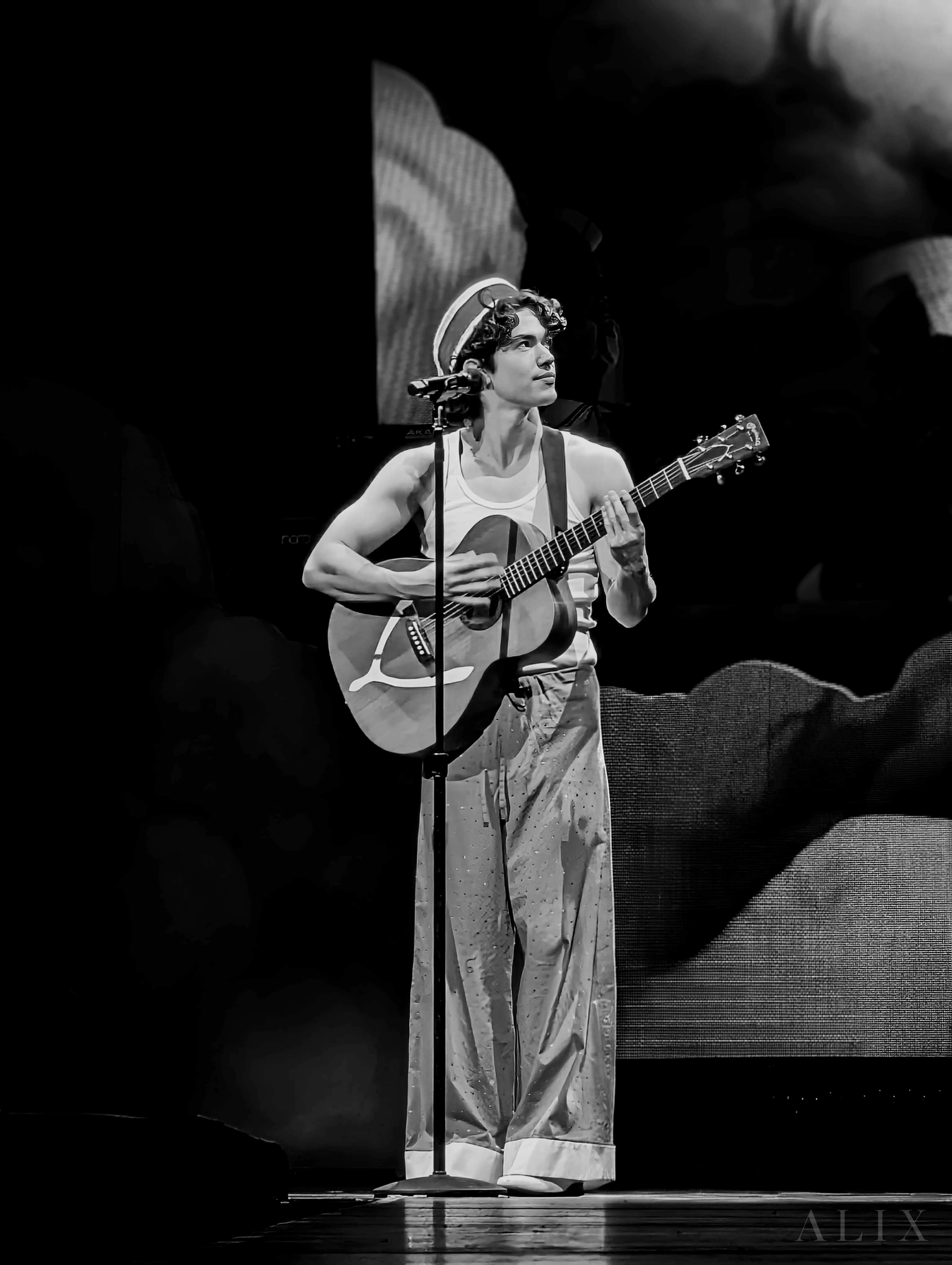 Black-and-white concert photograph of Conan Gray holding an acoustic guitar at a microphone.