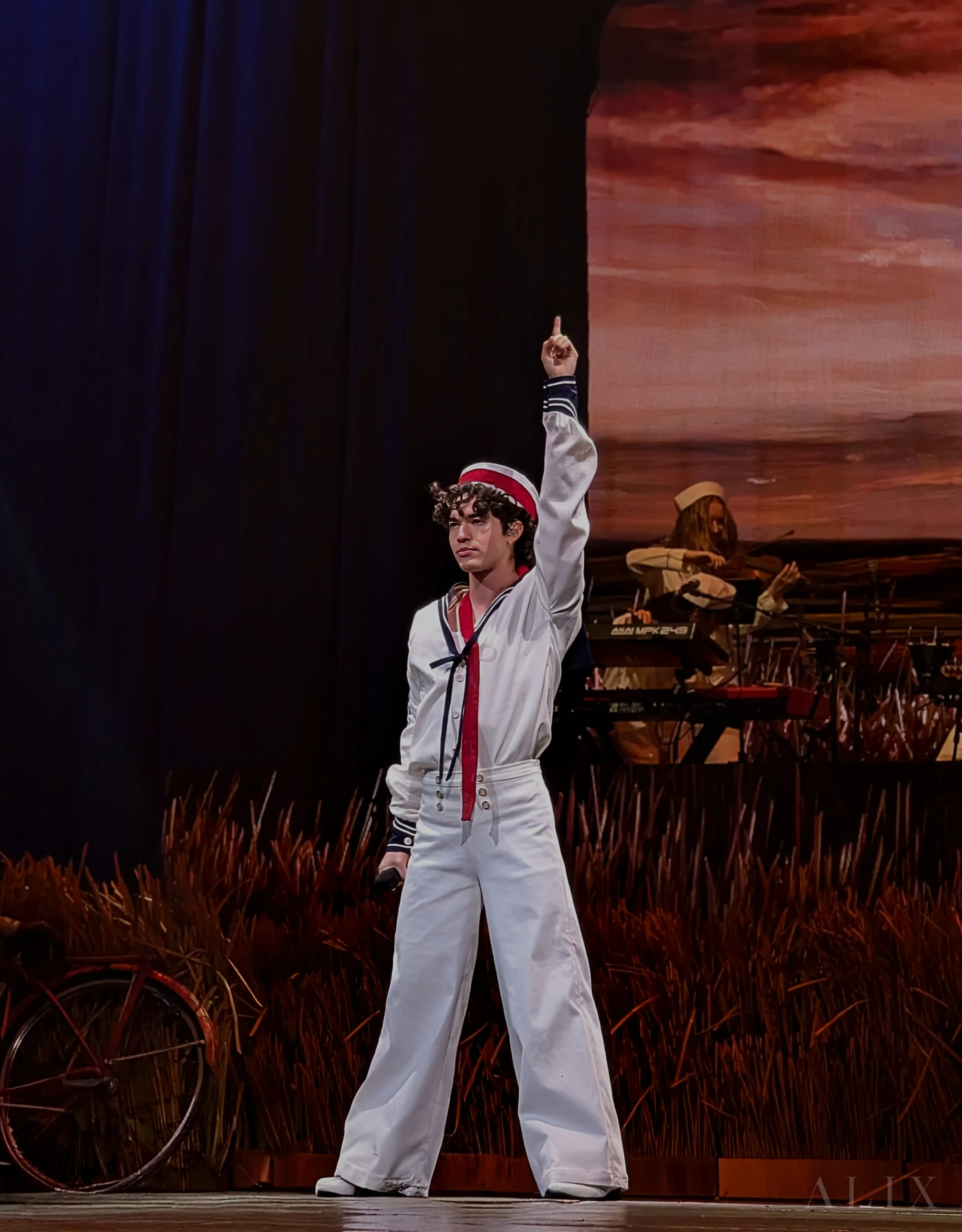 Conan Gray raises one arm onstage in a white sailor-inspired outfit against warm-colored scenery.