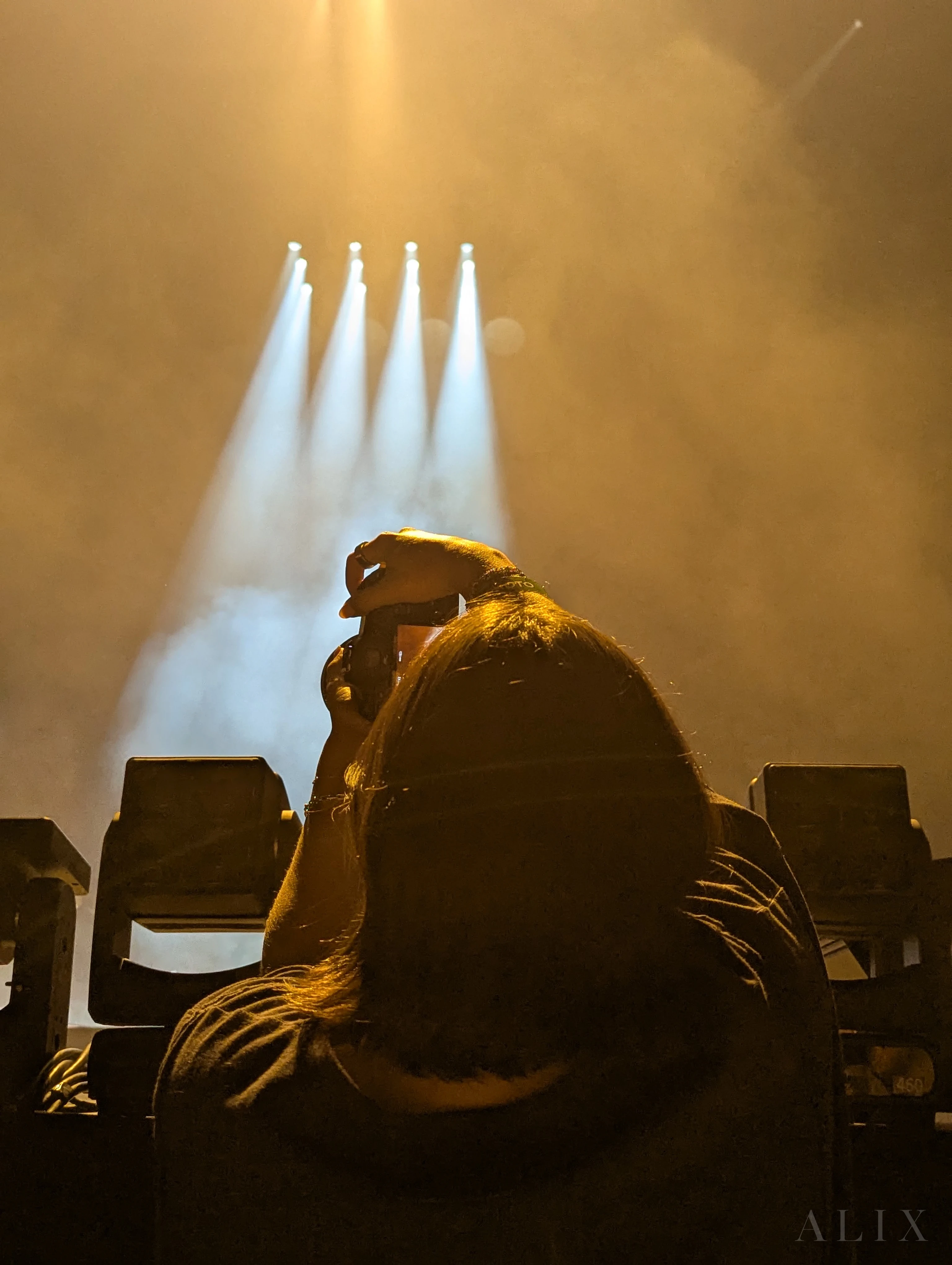 A concert photographer raises a camera beneath warm light and white spotlights.