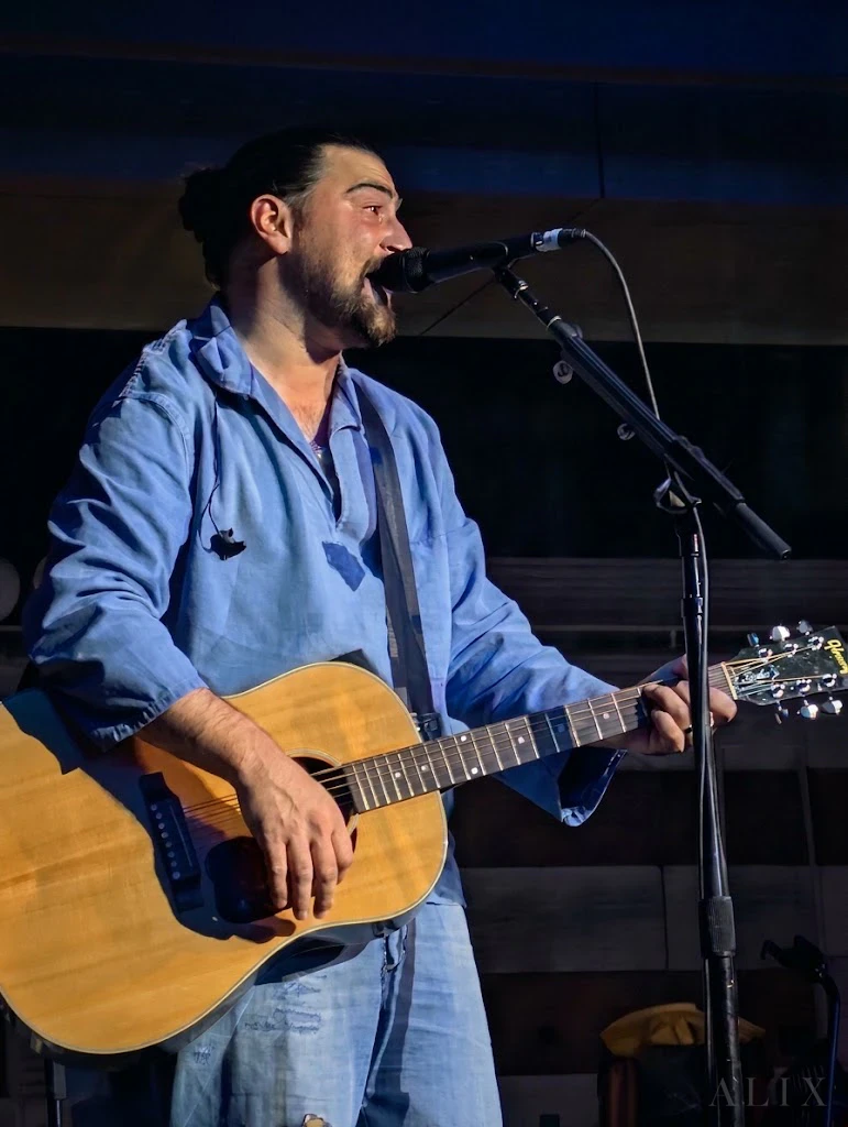 Noah Kahan sings into a microphone while holding an acoustic guitar, wearing a light-blue shirt.